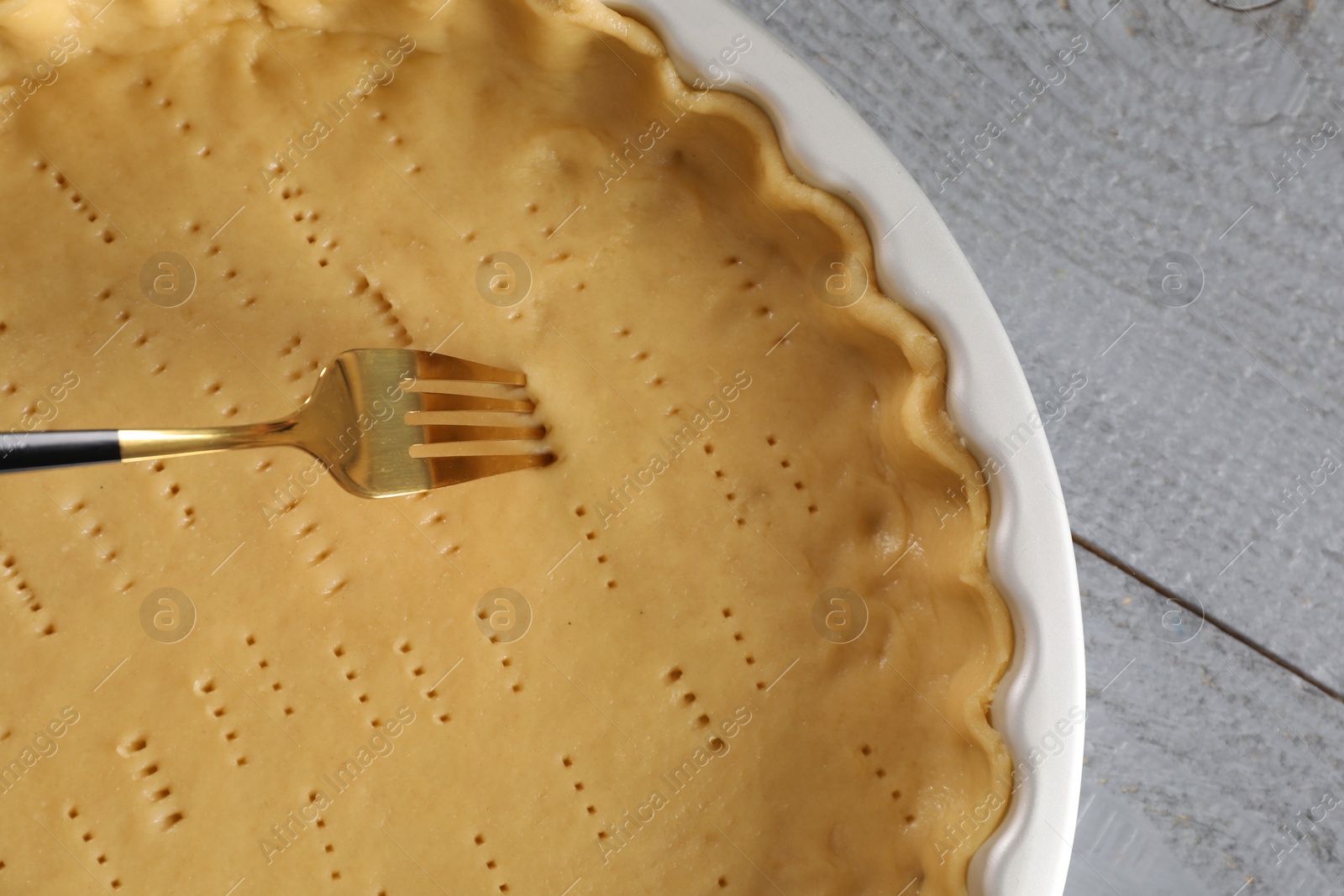 Photo of Pricking fresh dough with fork on grey wooden table, top view. Making quiche