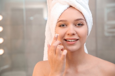 Beautiful woman with towel on head applying face cream in bathroom