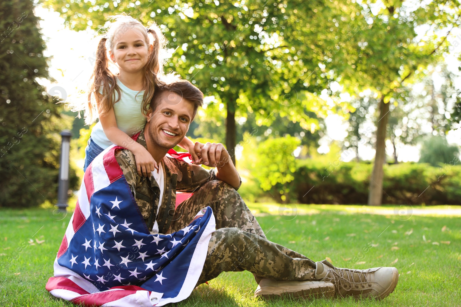 Photo of Father in military uniform with American flag and his little daughter sitting on grass at park