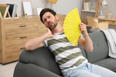 Bearded man waving yellow hand fan to cool himself on sofa at home