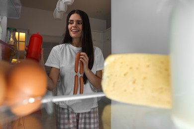 Young woman with sausages near modern refrigerator in kitchen at night, view from inside
