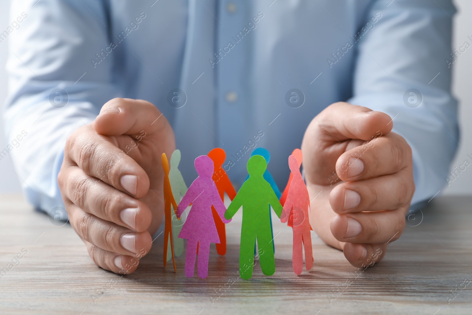 Photo of Man protecting paper human figures on wooden table, closeup. Diversity and inclusion concept