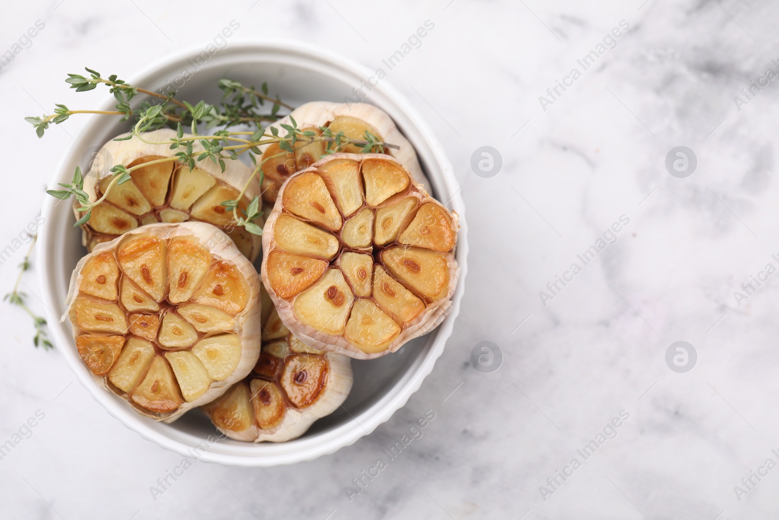 Photo of Heads of fried garlic and thyme in bowl on white table, top view. Space for text