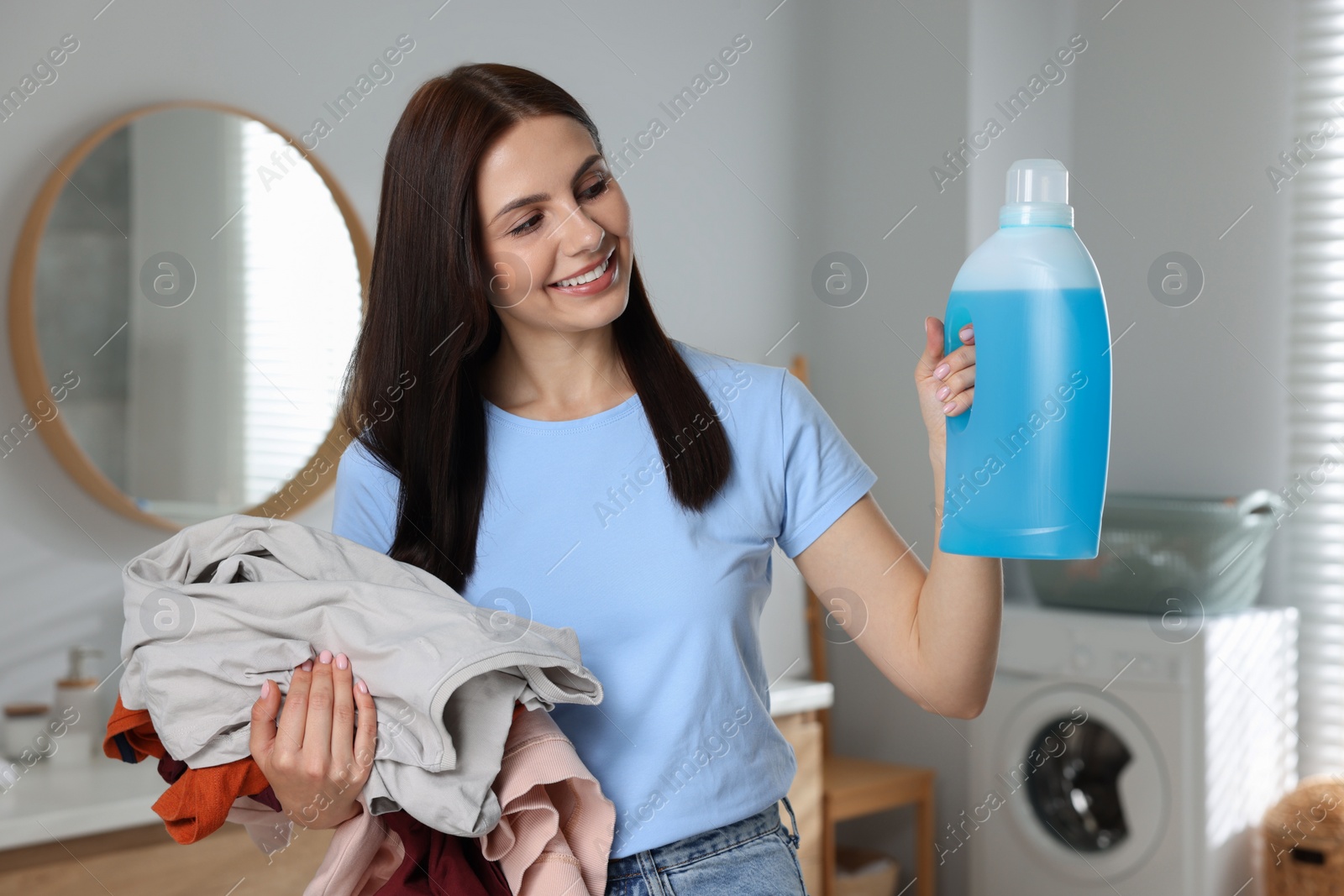 Photo of Woman holding fabric softener and dirty clothes in bathroom
