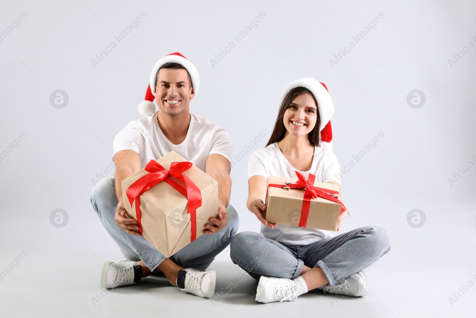 Photo of Beautiful happy couple in Santa hats sitting with Christmas gifts on light background