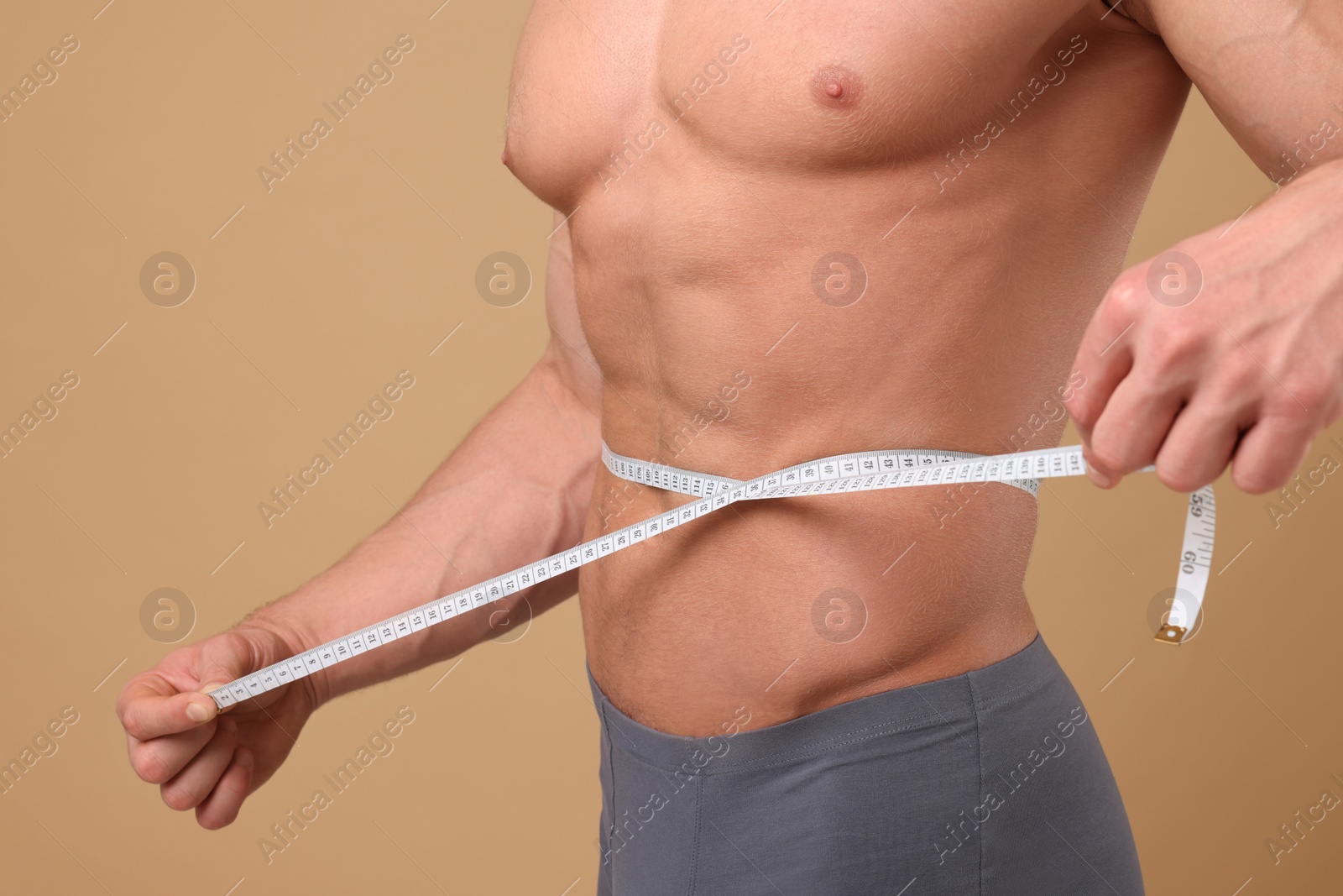 Photo of Athletic man measuring waist with tape on brown background, closeup. Weight loss concept