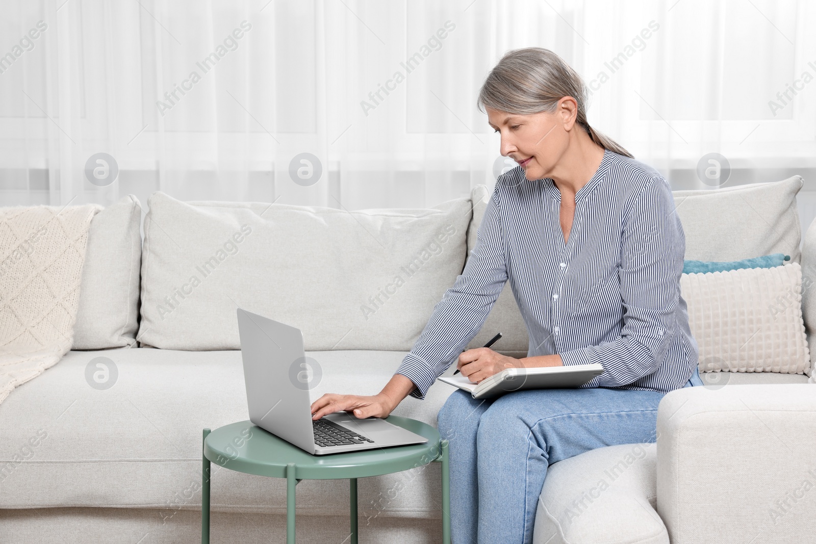 Photo of Beautiful senior woman writing something in notebook while using laptop at home