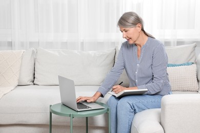 Photo of Beautiful senior woman writing something in notebook while using laptop at home