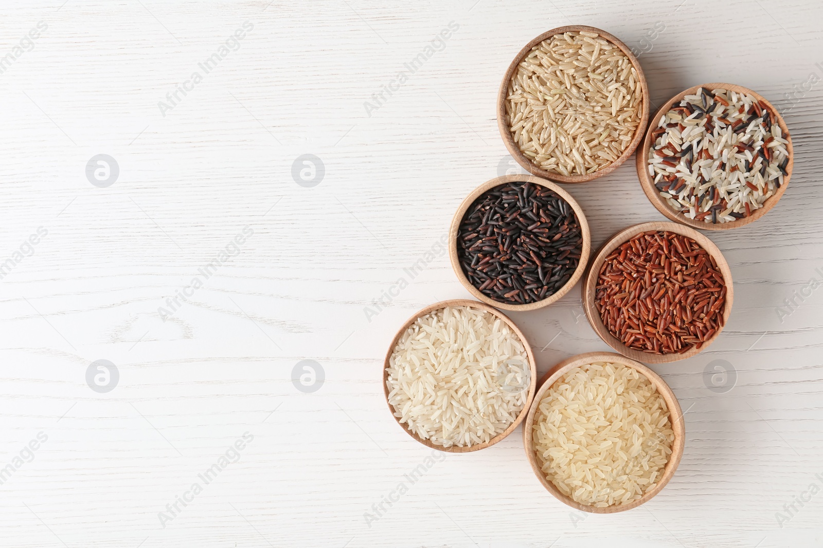 Photo of Flat lay composition with brown and other types of rice in bowls on white wooden background. Space for text