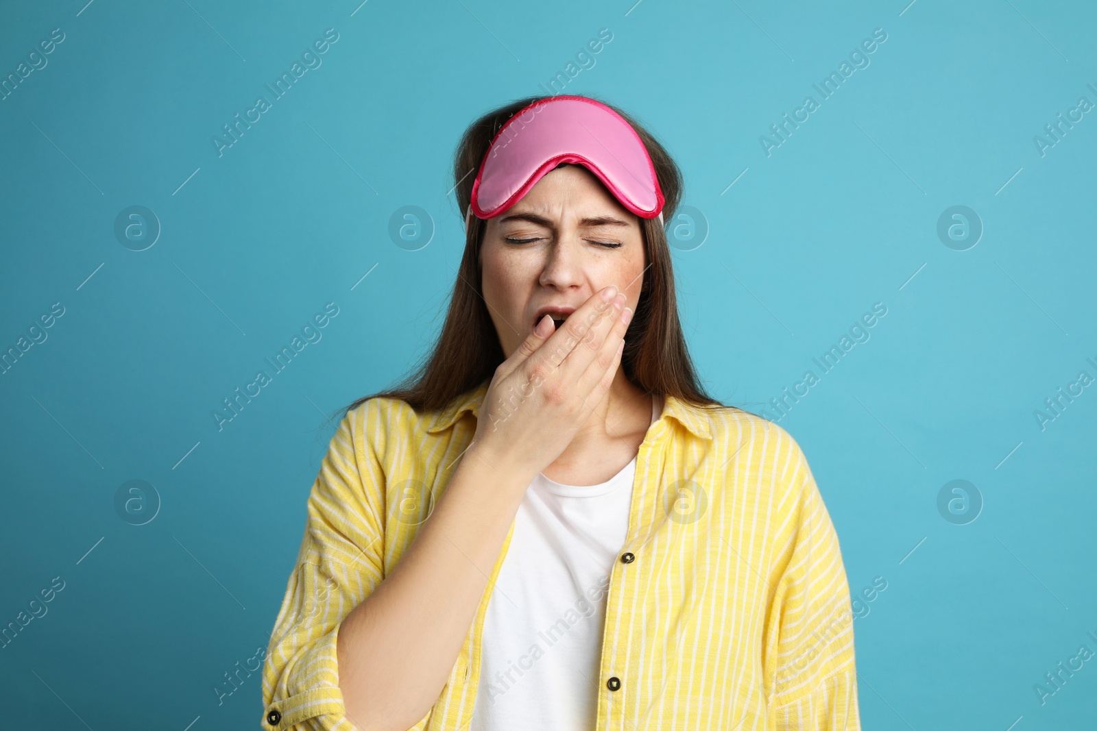 Photo of Young tired woman with sleeping mask yawning on light blue background