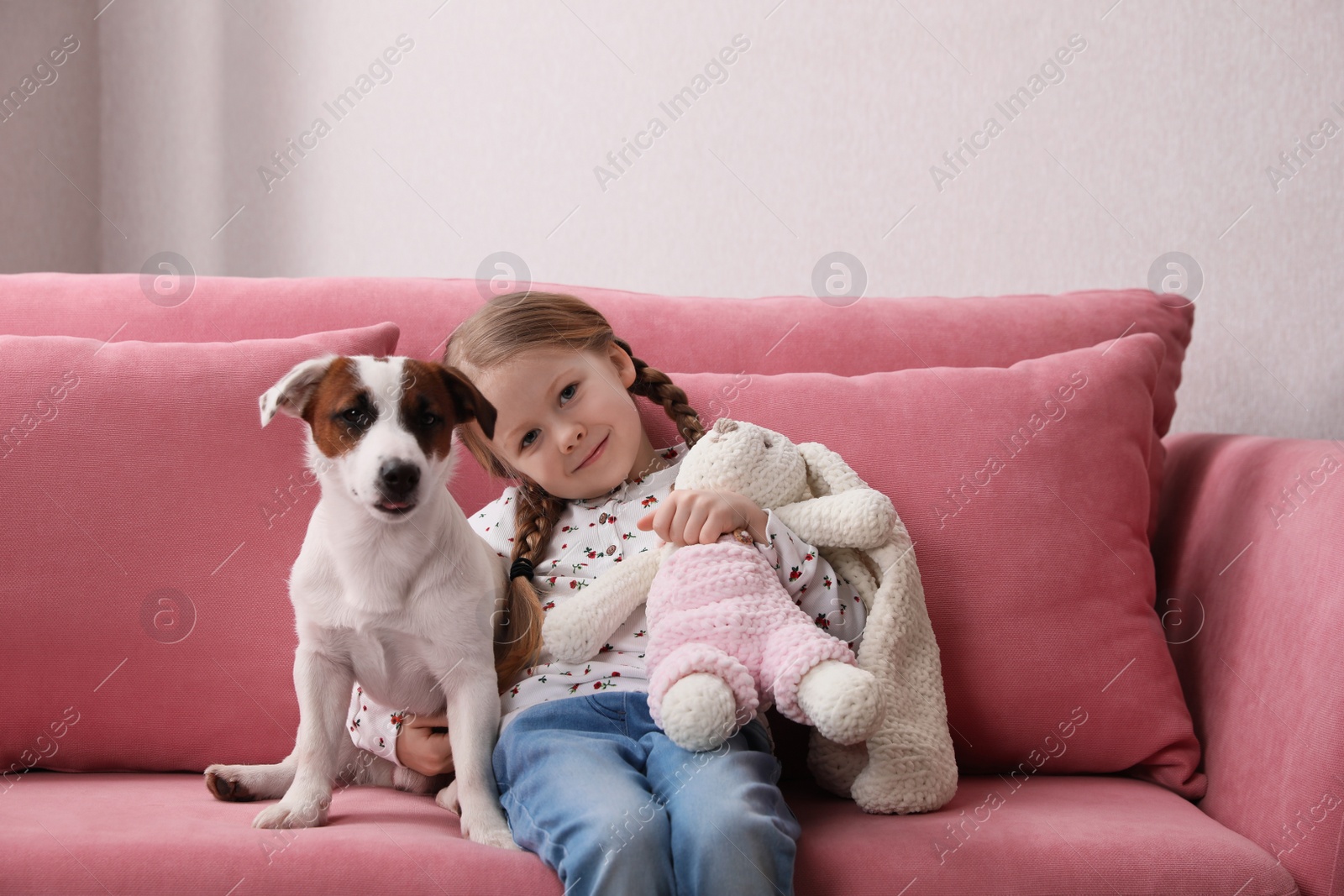 Photo of Cute little girl with her dog and toy bunny on sofa indoors. Childhood pet