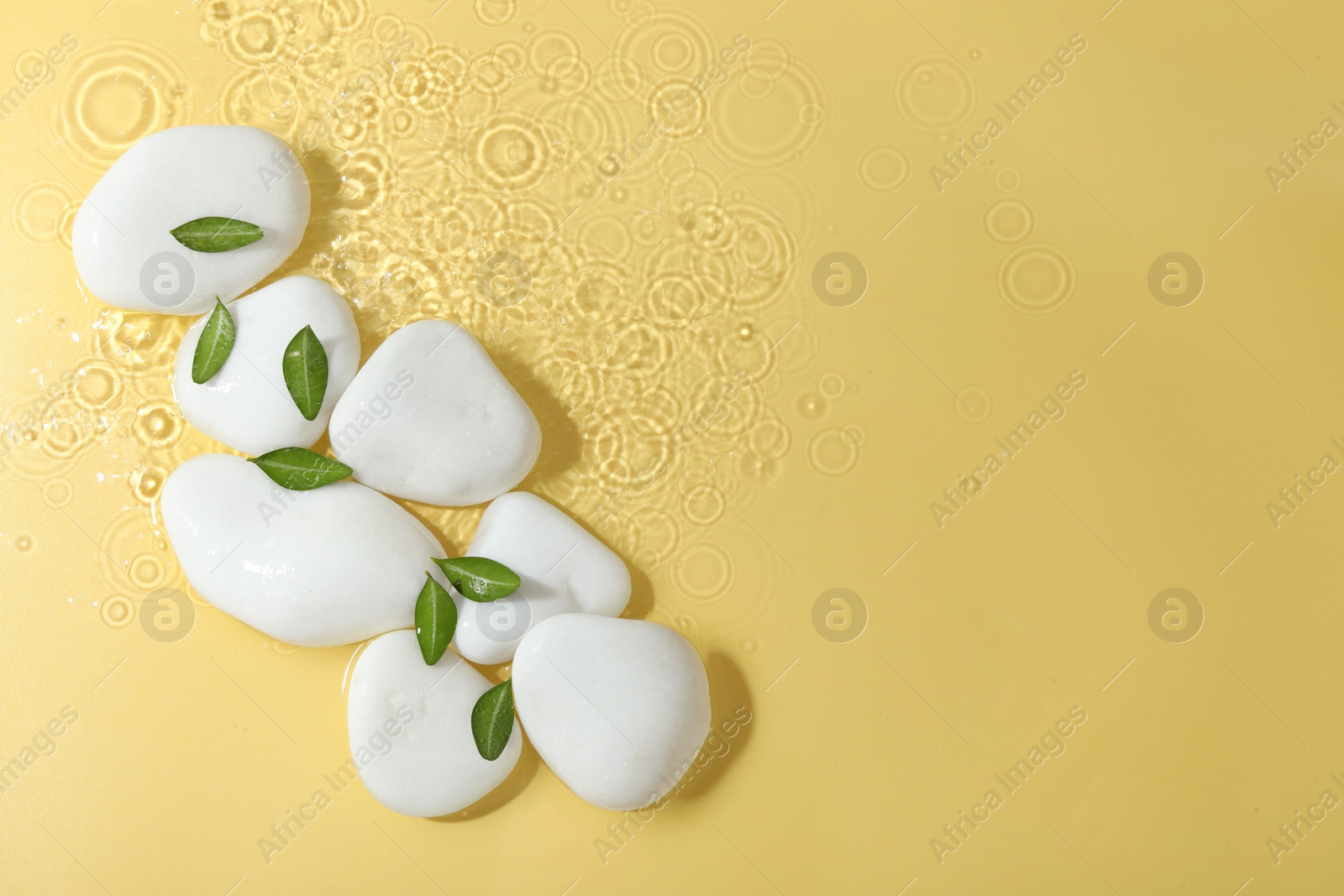 Photo of Spa stones and green leaves in water on yellow background, flat lay. Space for text