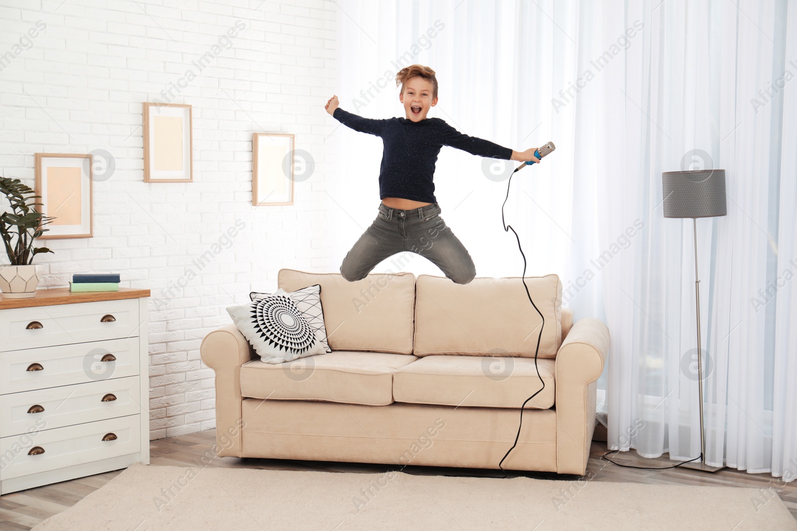 Photo of Cute boy with microphone jumping on sofa in living room