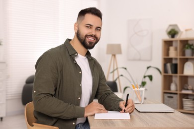 Photo of Young man writing in notebook at wooden table indoors