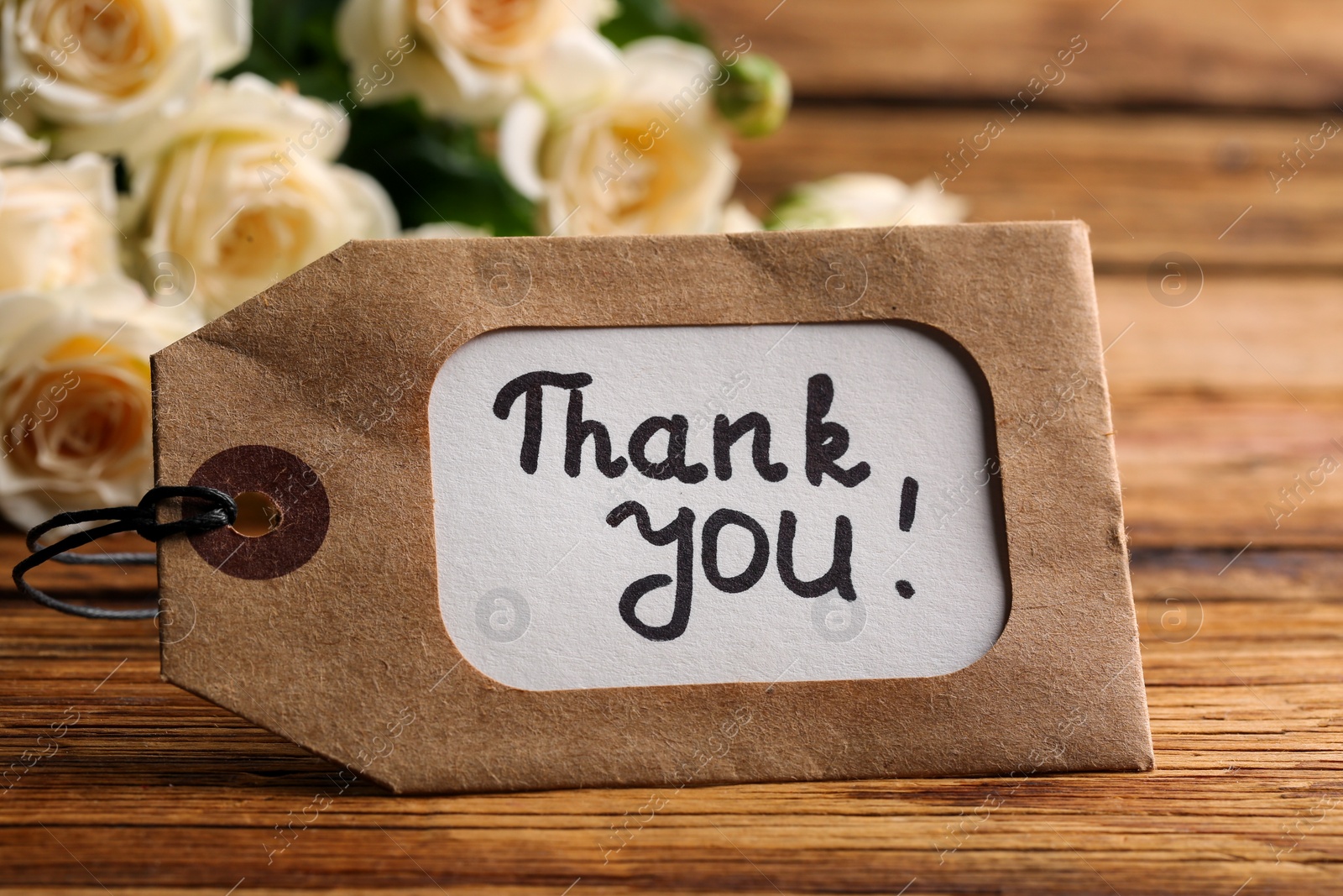 Photo of Tag with phrase Thank you and roses on wooden table, closeup