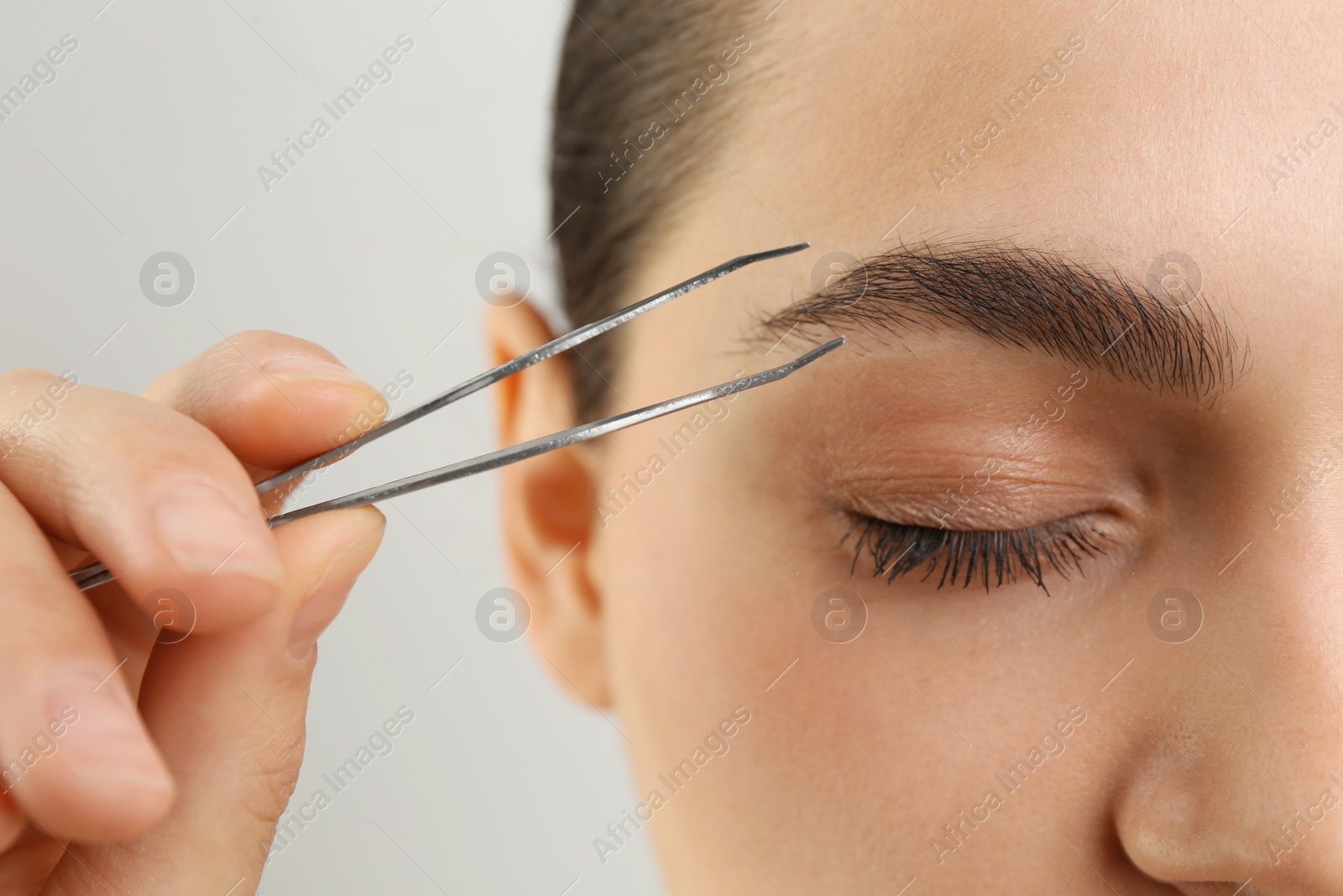 Photo of Eyebrow correction. Young woman with tweezers on light grey background, closeup