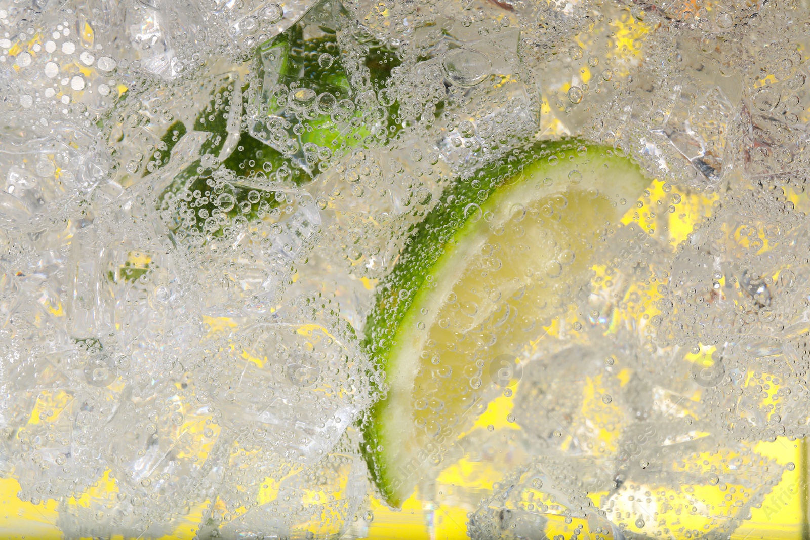 Photo of Juicy lime slices and ice cubes in soda water against yellow background, closeup