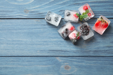 Ice cubes with different berries and mint on blue wooden table, flat lay. Space for text
