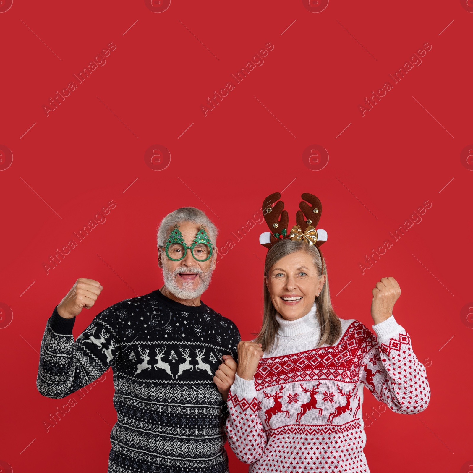 Photo of Happy senior couple in Christmas sweaters, reindeer headband and funny glasses on red background