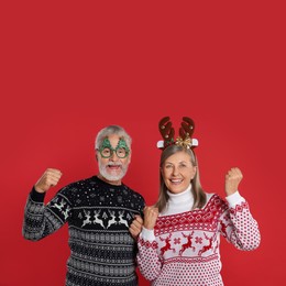 Happy senior couple in Christmas sweaters, reindeer headband and funny glasses on red background