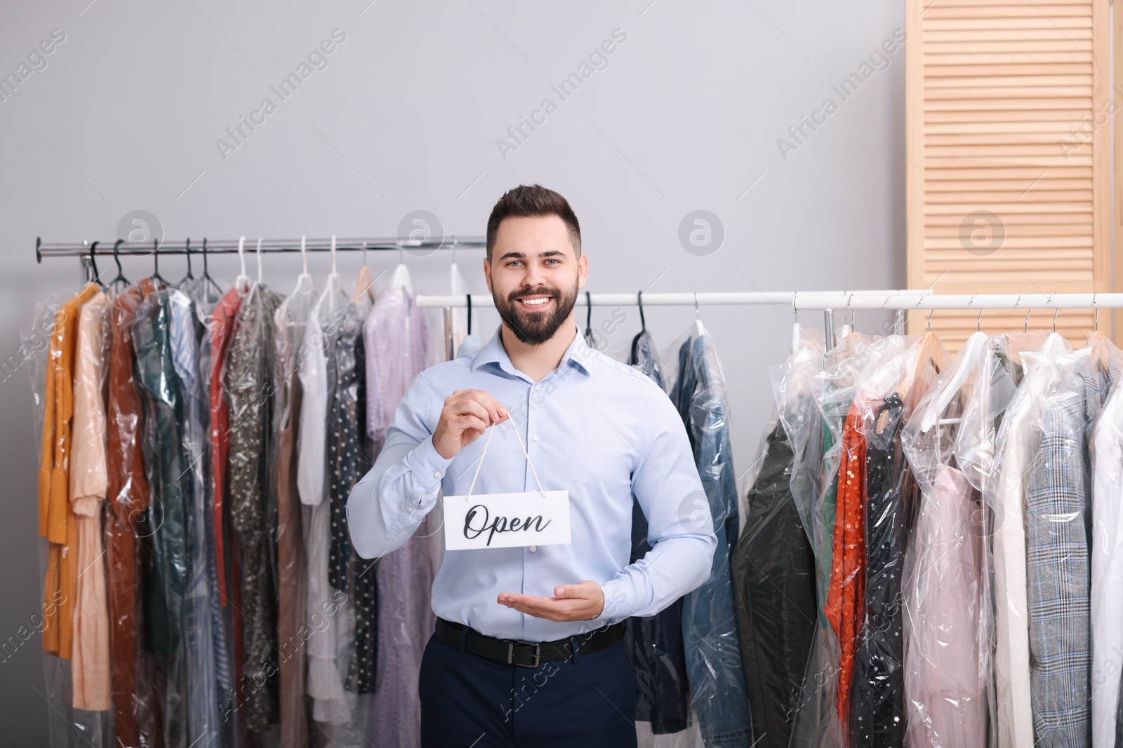 Photo of Dry-cleaning service. Happy worker holding Open sign near racks with clothes indoors