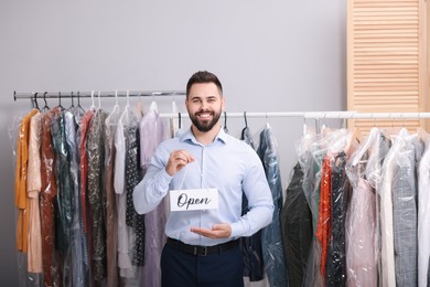 Dry-cleaning service. Happy worker holding Open sign near racks with clothes indoors