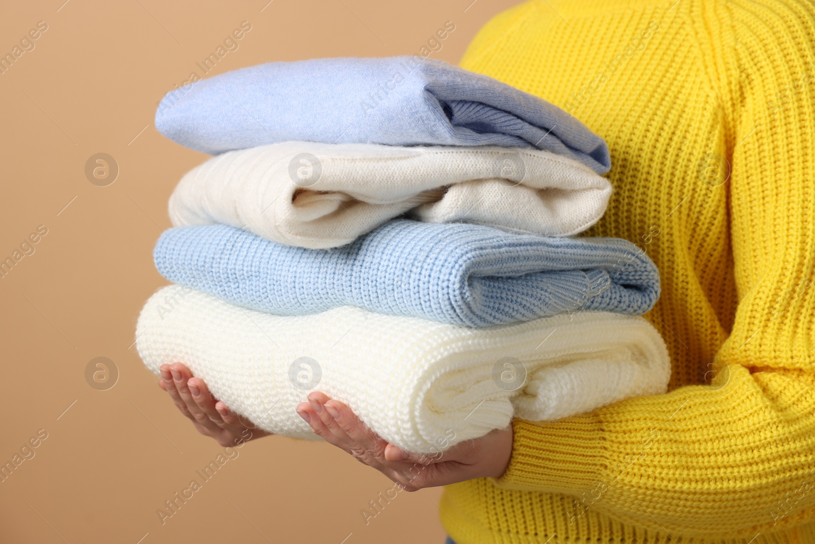 Photo of Woman with stack of casual sweaters on light brown background, closeup