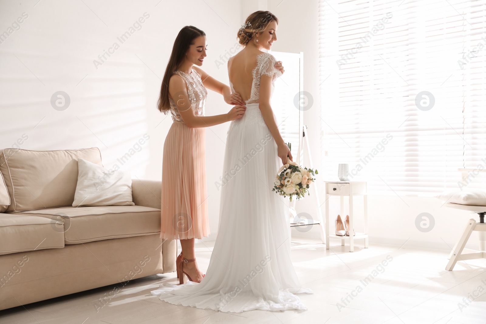 Photo of Young woman helping bride to put on wedding dress in room