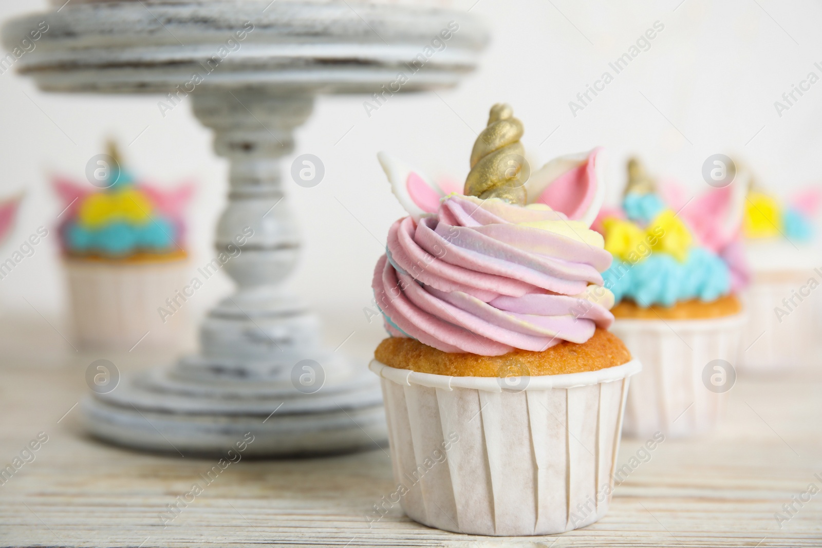 Photo of Cute sweet unicorn cupcake near dessert stand on white wooden table, closeup