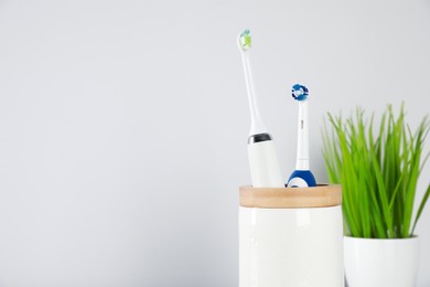 Photo of Electric toothbrushes in holder and green houseplant on light background. Space for text