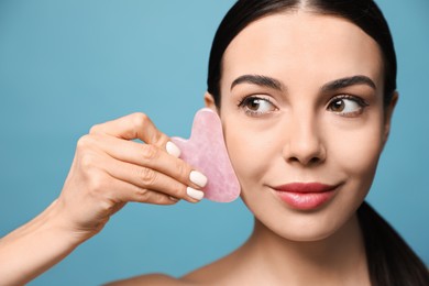 Photo of Beautiful young woman doing facial massage with gua sha tool on blue background, closeup