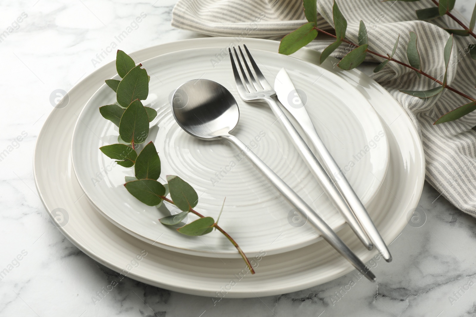 Photo of Stylish setting with cutlery, napkin, eucalyptus branches and plates on white marble table, closeup