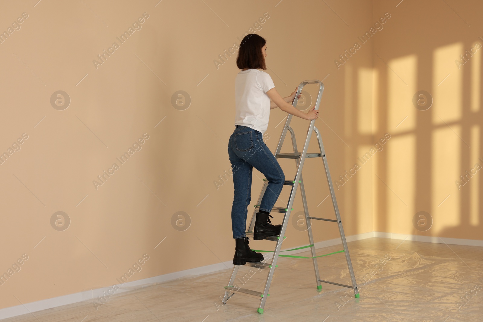 Photo of Young woman climbing up stepladder near wall indoors. Room renovation