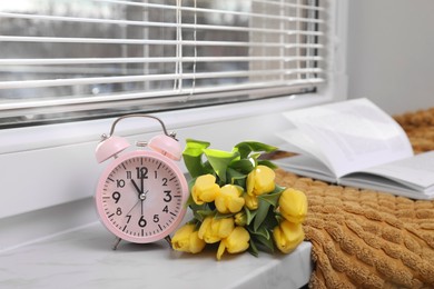 Photo of Pink alarm clock, soft blanket, book and wonderful tulips on window sill indoors. Spring atmosphere