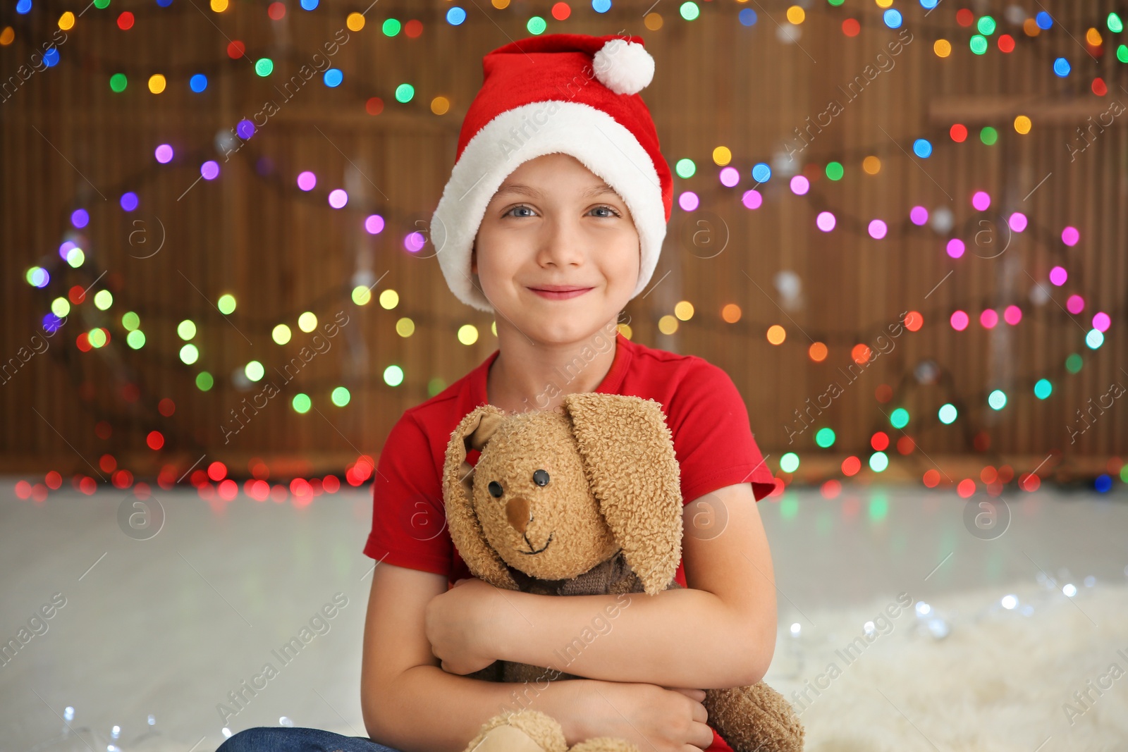 Photo of Cute little child in Santa hat with toy rabbit on blurred lights background. Christmas celebration