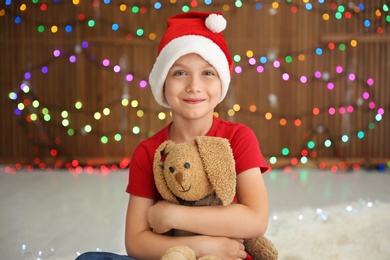 Cute little child in Santa hat with toy rabbit on blurred lights background. Christmas celebration