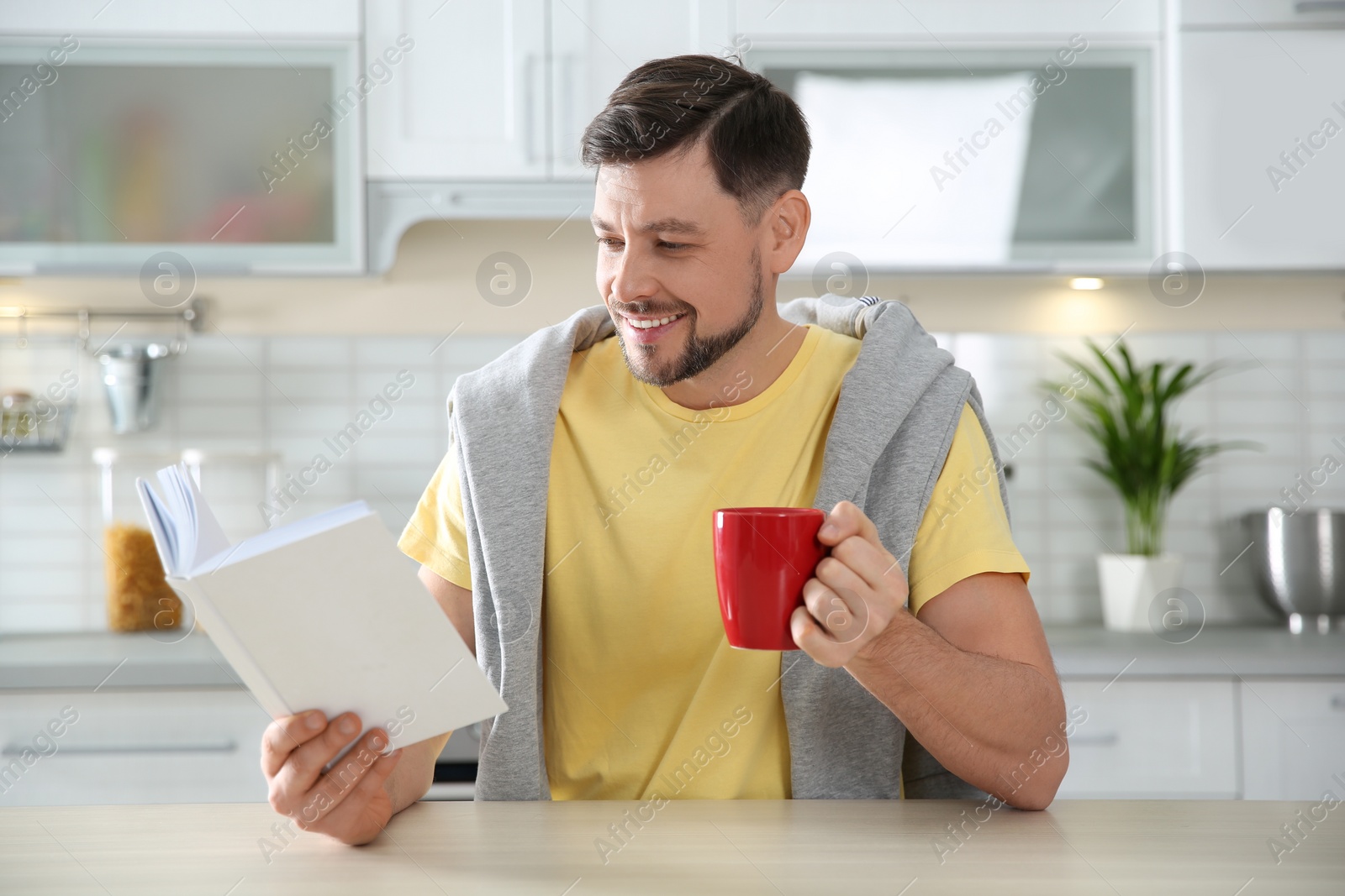 Photo of Handsome man with cup of coffee reading book at table in kitchen