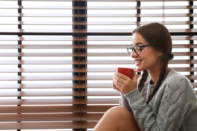 Image of Happy young woman with cup of hot drink near window. Lazy morning