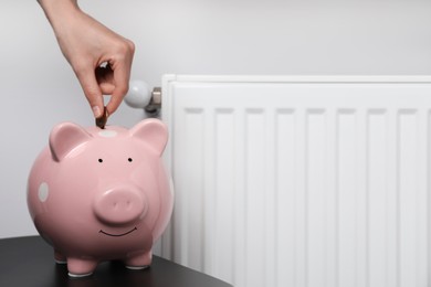 Photo of Woman putting coin into piggy bank near heating radiator, closeup. Space for text