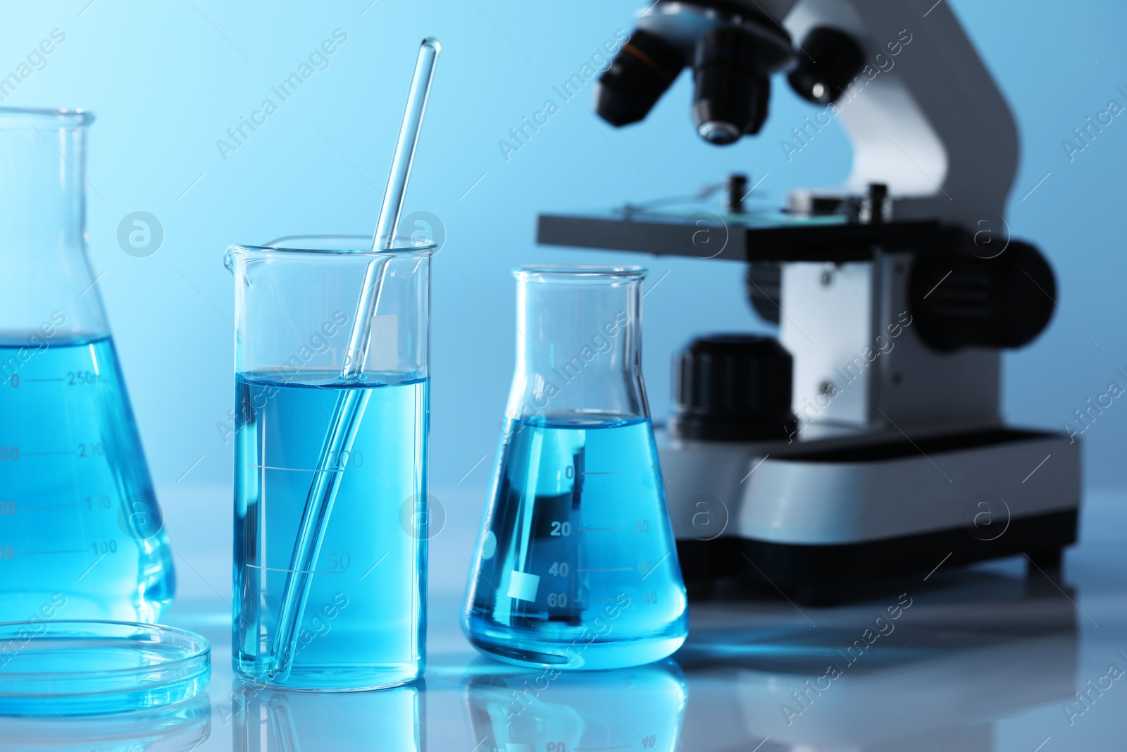 Photo of Different laboratory glassware with light blue liquid and microscope on table