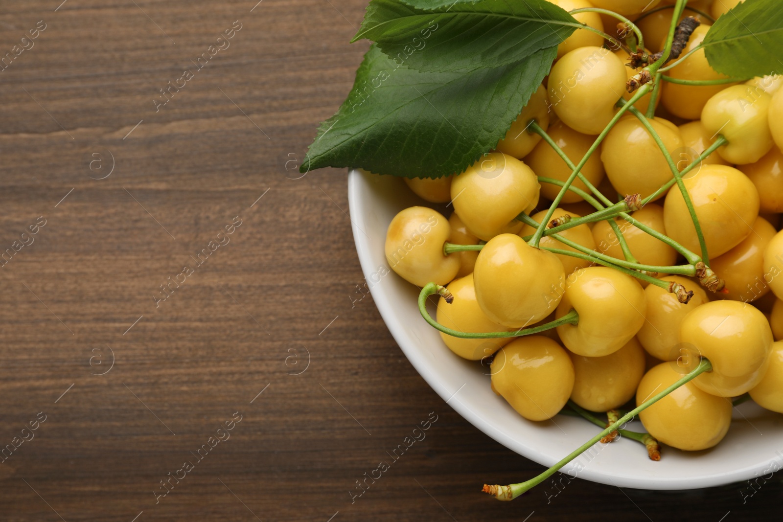 Photo of Bowl with ripe yellow cherries and green leaves on wooden table, top view. Space for text