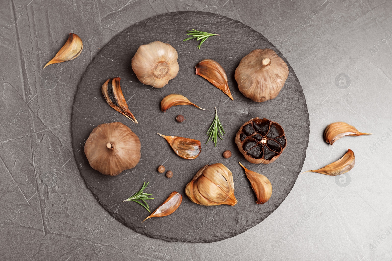 Photo of Slate plate with aged black garlic and rosemary on gray background, top view