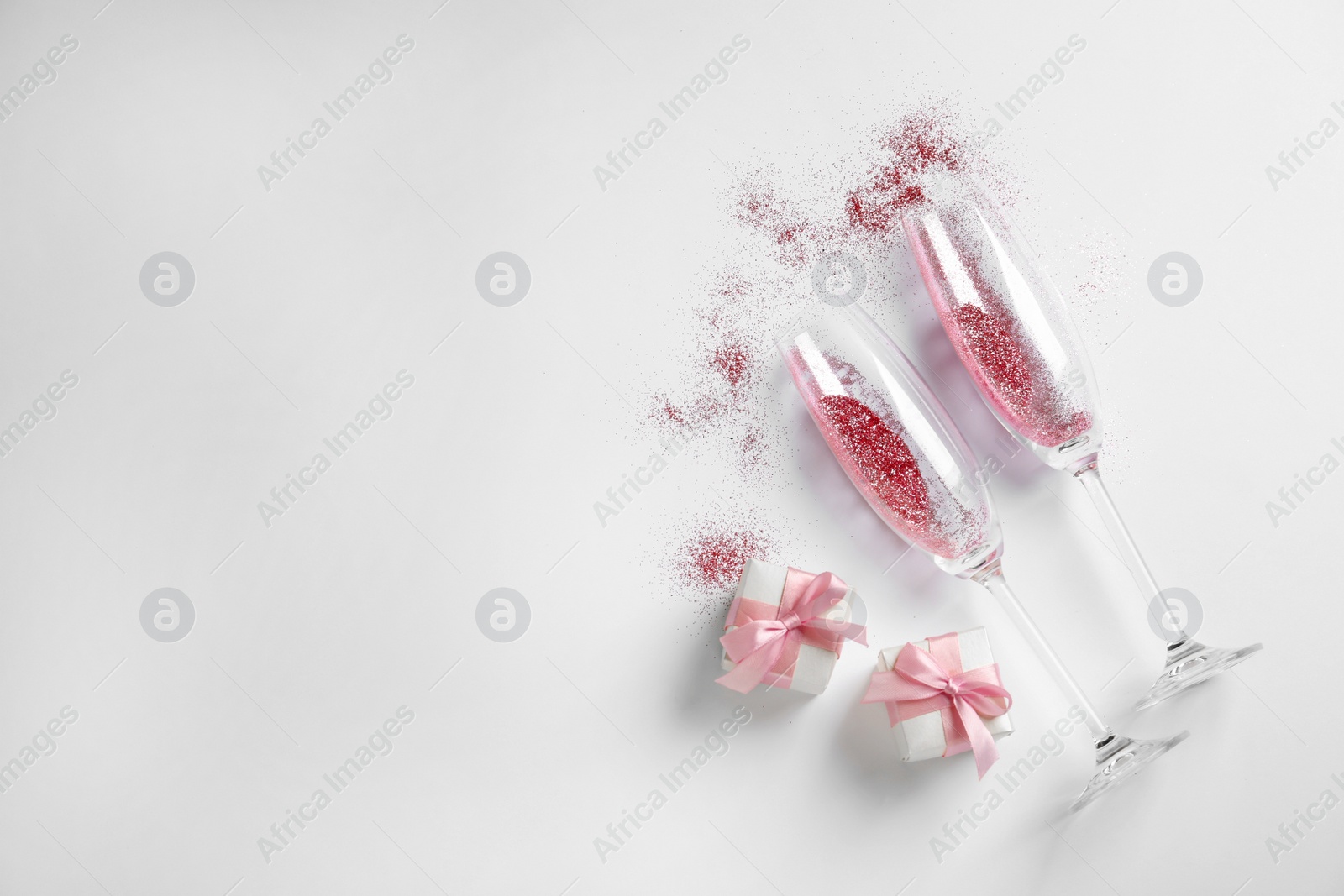 Photo of Champagne glasses with pink glitter, gift boxes and space for text on white background, top view. Hilarious celebration