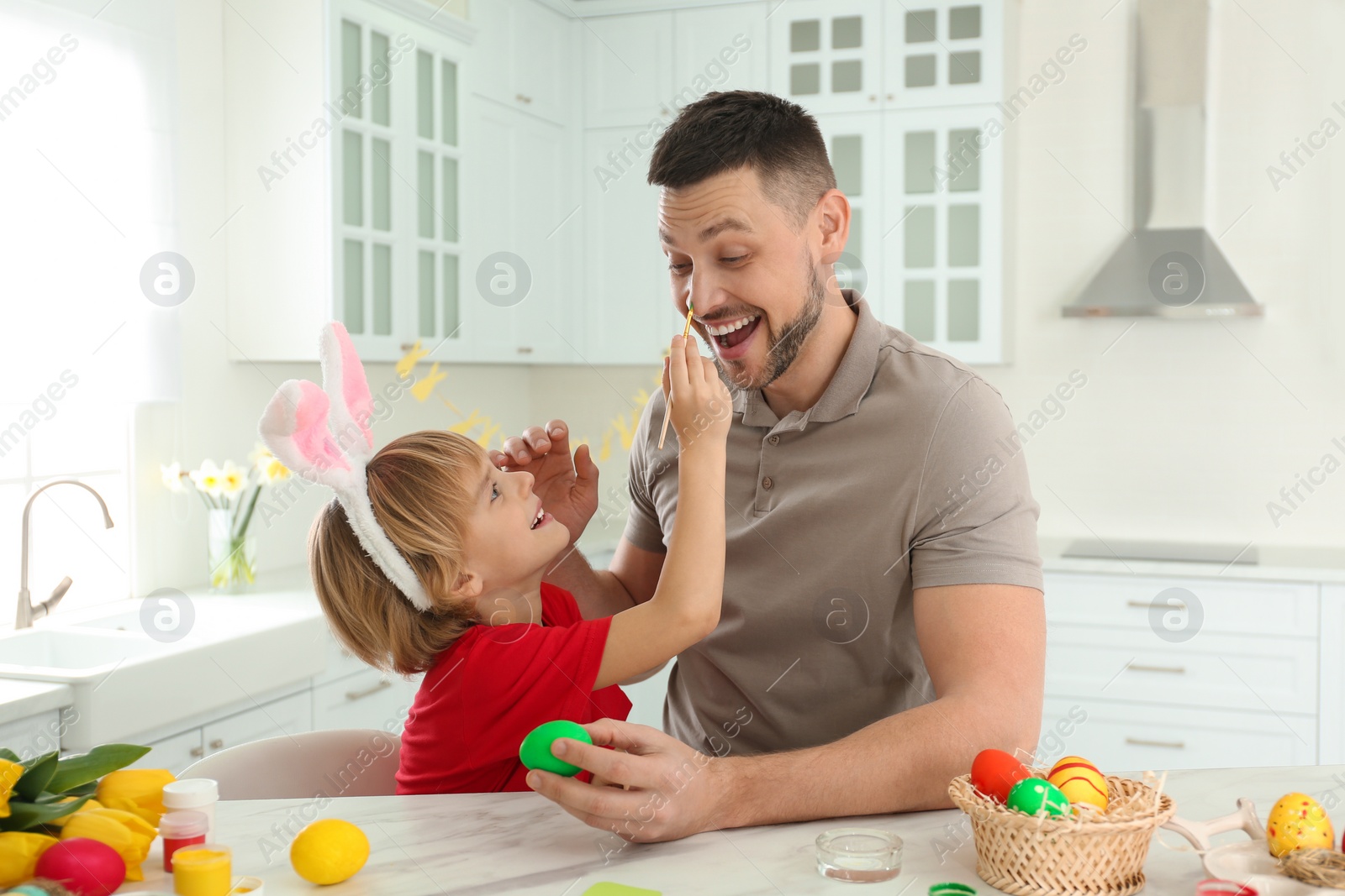 Photo of Happy son with bunny ears headband and his father having fun while painting Easter egg at table in kitchen