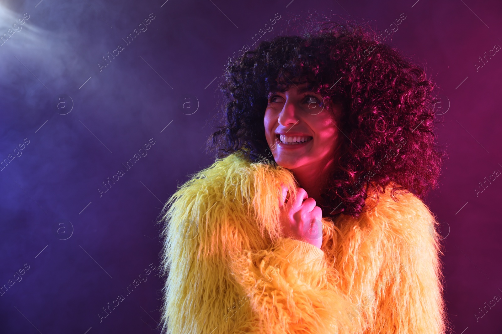 Photo of Beautiful young woman in yellow fur coat on color background in neon lights and smoke