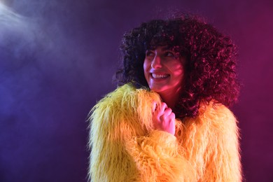 Photo of Beautiful young woman in yellow fur coat on color background in neon lights and smoke