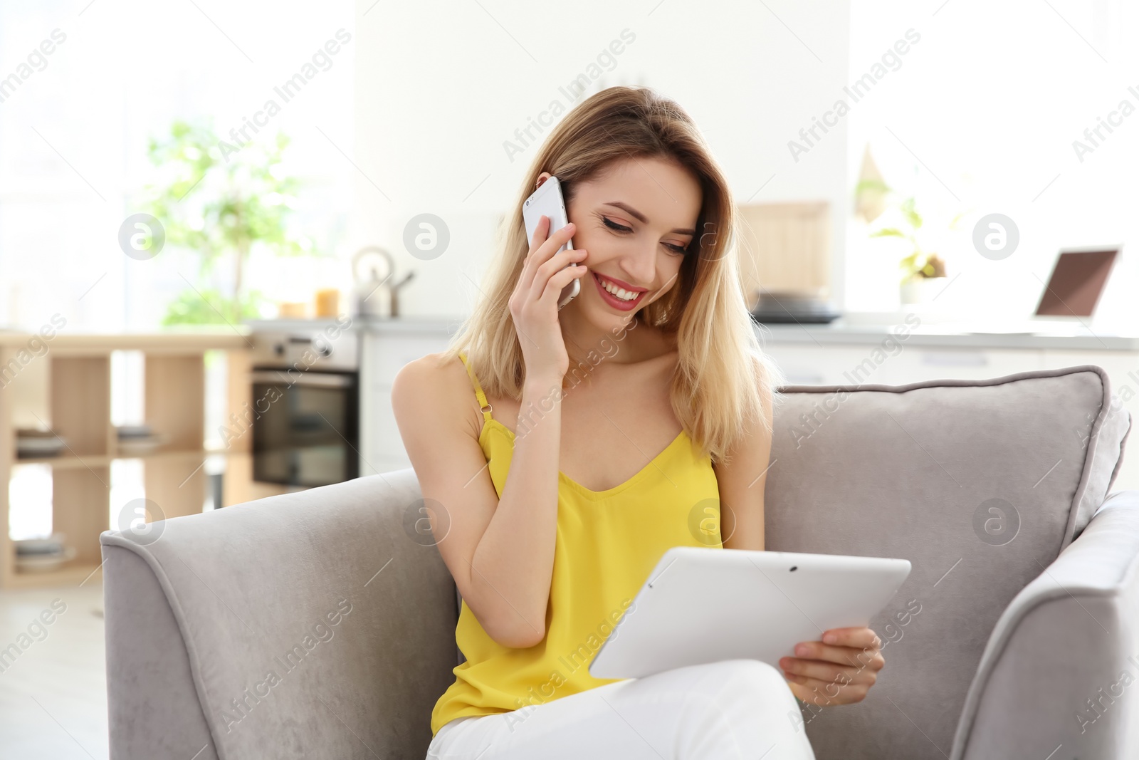 Photo of Young woman talking on phone while using tablet indoors