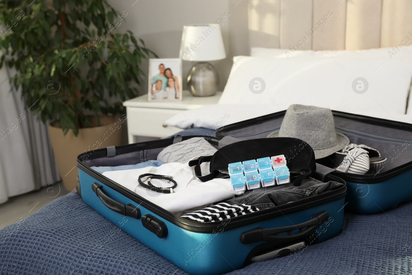 Photo of Open suitcase with packed clothes, accessories and pill box on bed indoors