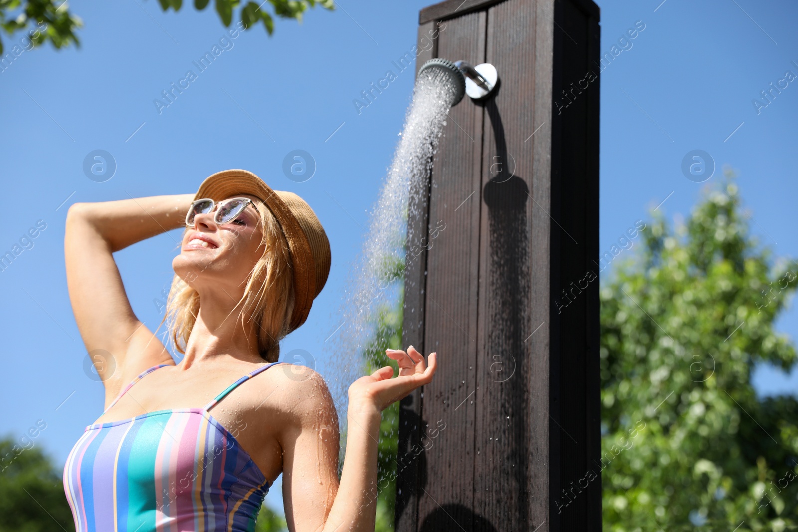 Photo of Sexy young woman in stylish bikini with hat and sunglasses taking shower outdoors on sunny day