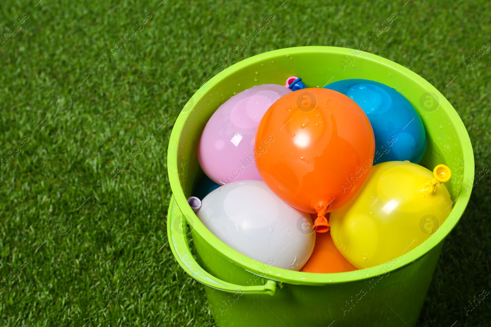 Photo of Bucket full of colorful water bombs on green grass, closeup. Space for text